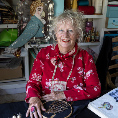 An image of Susie Vickery at a table, with a partially completed cross stitch on the table in front of her.
